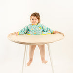 Bavoir de rechange bleu-vert à motifs pour kits BibaTray M et L, vue complète sur bébé. | Replacement blue-green patterned bib for BibaTray M and L kits, full view on baby.