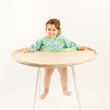 Bavoir de rechange bleu-vert à motifs pour kits BibaTray M et L, vue complète sur bébé. | Replacement blue-green patterned bib for BibaTray M and L kits, full view on baby.