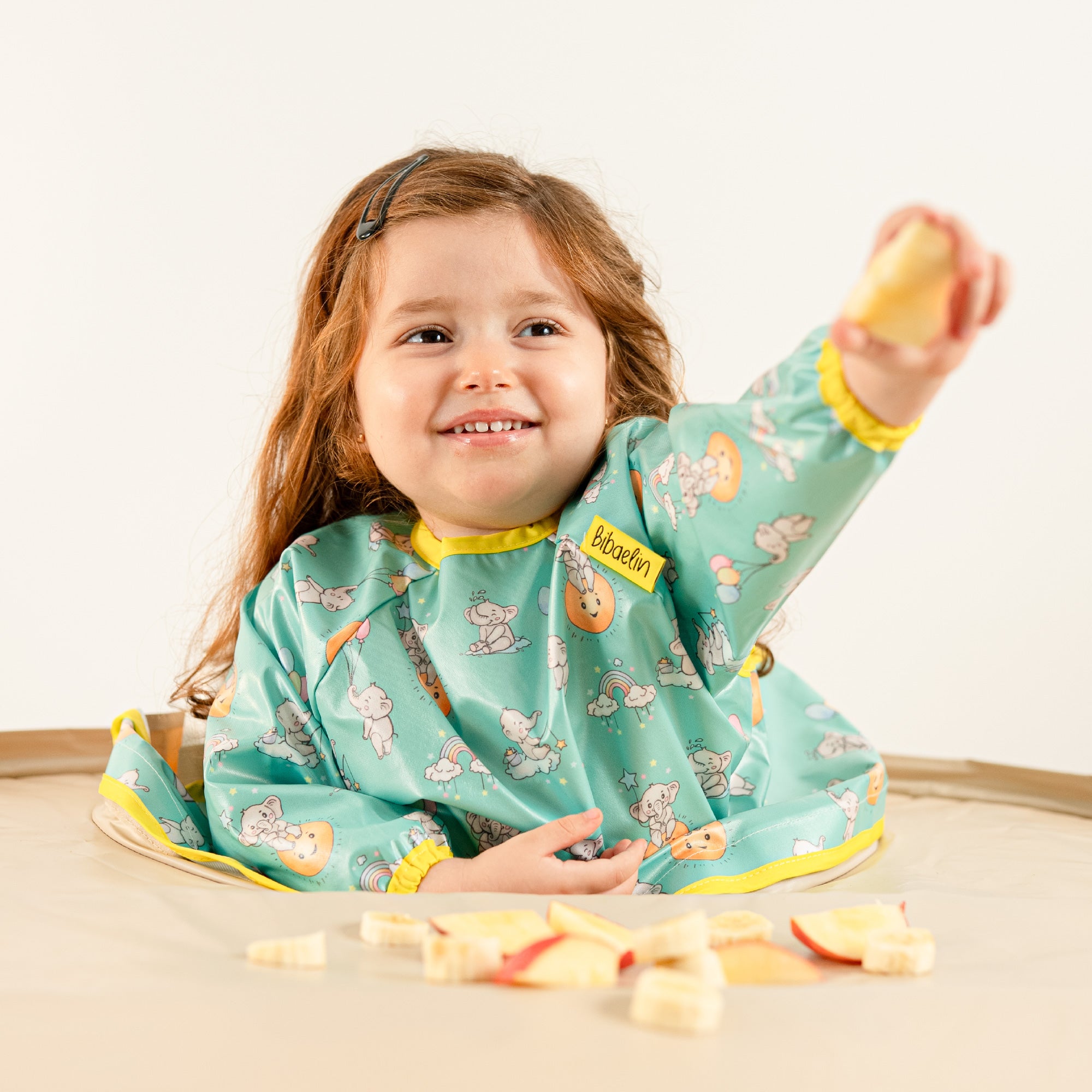 Bébé mangeant des morceaux de pomme en toute autonomie avec le bavoir bleu-vert Bibaelin. | Baby self-feeding apple pieces with the teal Bibaelin bib.
