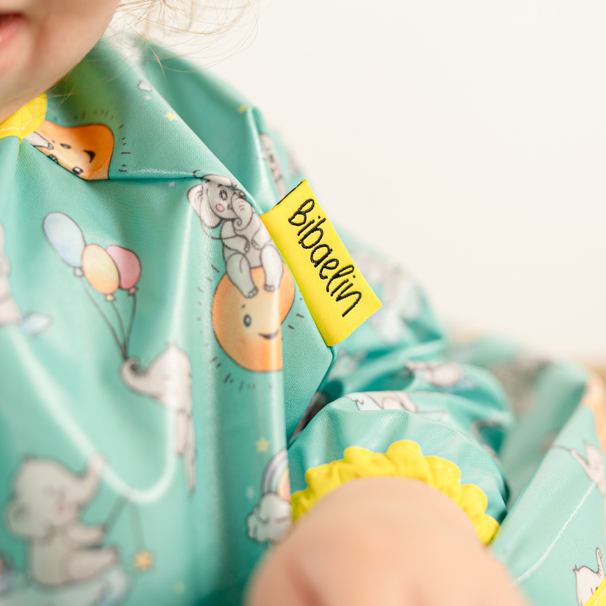 Gros plan sur l'étiquette jaune Bibaelin et les motifs éléphants du bavoir bleu-vert - Close-up | Close-up of the yellow Bibaelin label and elephant patterns of the teal blue-green bib