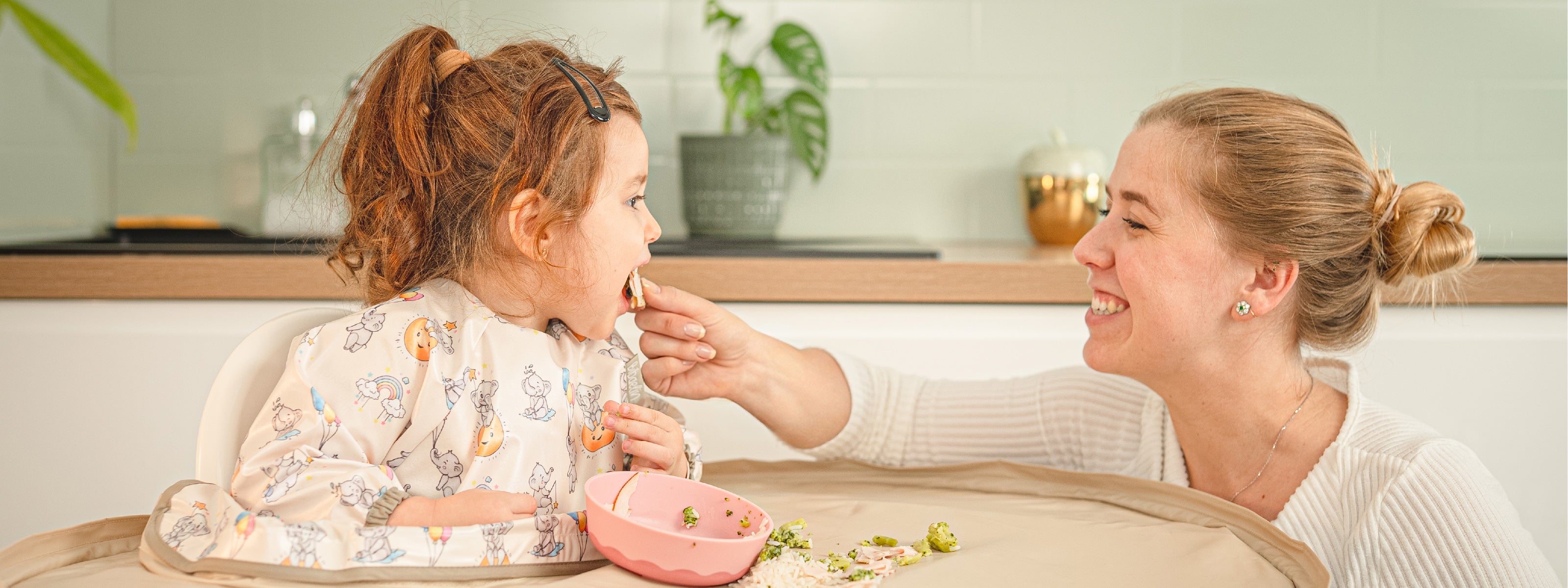 Mère souriante aidant son enfant à manger avec le bavoir plateau intégral Bibaelin taille L pendant un repas de diversification - Mother smiling and feeding her toddler with the large Bibaelin bib tray kit.